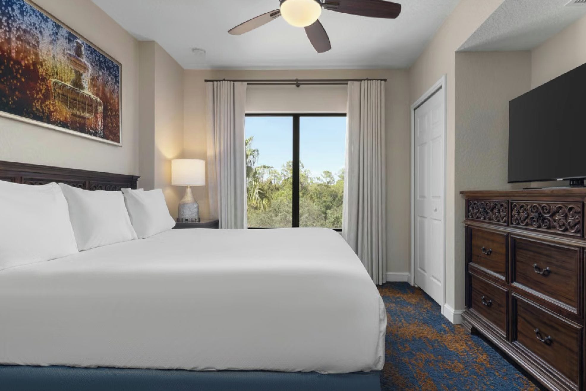 A hotel room with a large bed, a ceiling fan, a television, and a view of the outdoors.