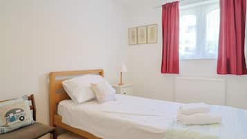 Una cama individual con ropa blanca, una mesita de noche con lámpara, un sillón con cojín y una ventana con cortinas rojas.