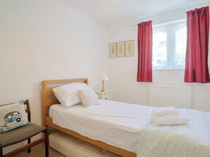 Una cama individual con ropa blanca, una mesita de noche con lámpara, un sillón con cojín y una ventana con cortinas rojas.