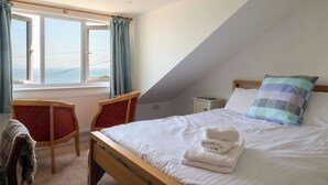 A bedroom with a bed, a desk, two chairs, a window with curtains, and a view of the sea. - Seaview (Lyme Regis)