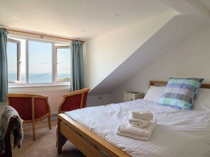 Ein Schlafzimmer mit einem Bett, einem Schreibtisch, zwei Stühlen, einem Fenster mit Vorhängen und Blick aufs Meer.