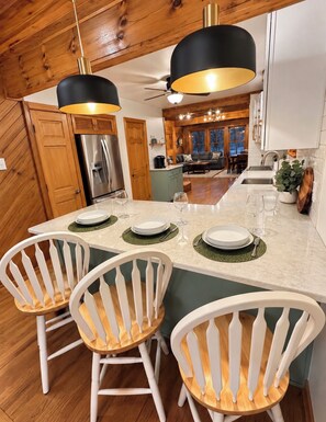 A modern kitchen with wooden cabinets, a stainless steel refrigerator, and a dining area with white chairs and a marble countertop. - Great Escape at Lake Harmony • 4BR • Beach Access • Game Room • Fire Pit (Lake Harmony)