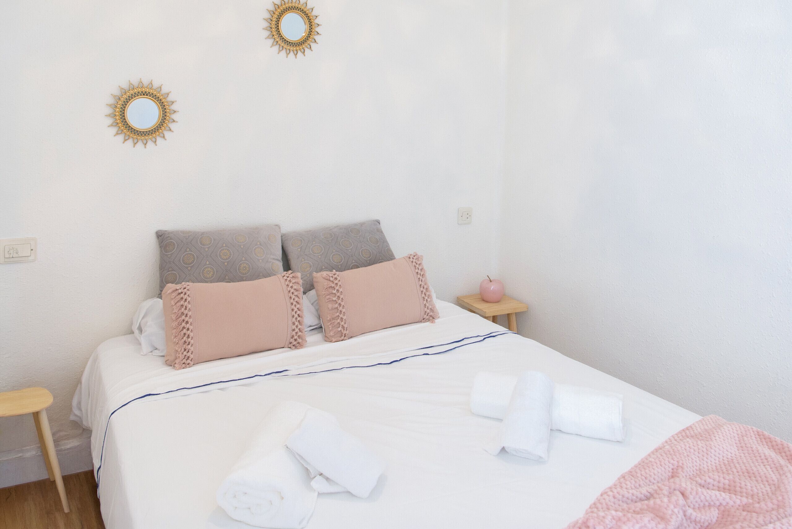 Una cama bien hecha con almohadas, una mesita de noche con una lámpara rosa y un espejo en la pared.