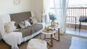 Ein gemütliches Wohnzimmer mit einer Couch, zwei kleinen Tischen mit Pflanzen, einem Traumfänger und einem Balkon mit Sitzgelegenheiten.