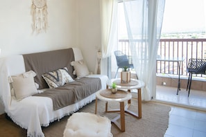 Een gezellige woonkamer met een bank, twee bijzettafeltjes met planten, een dromenvanger en een balkon met stoelen.