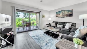 Un salon moderne avec un canapé, une table basse et un téléviseur. Une porte-fenêtre coulissante donne accès à un espace extérieur aménagé avec une table et des chaises.