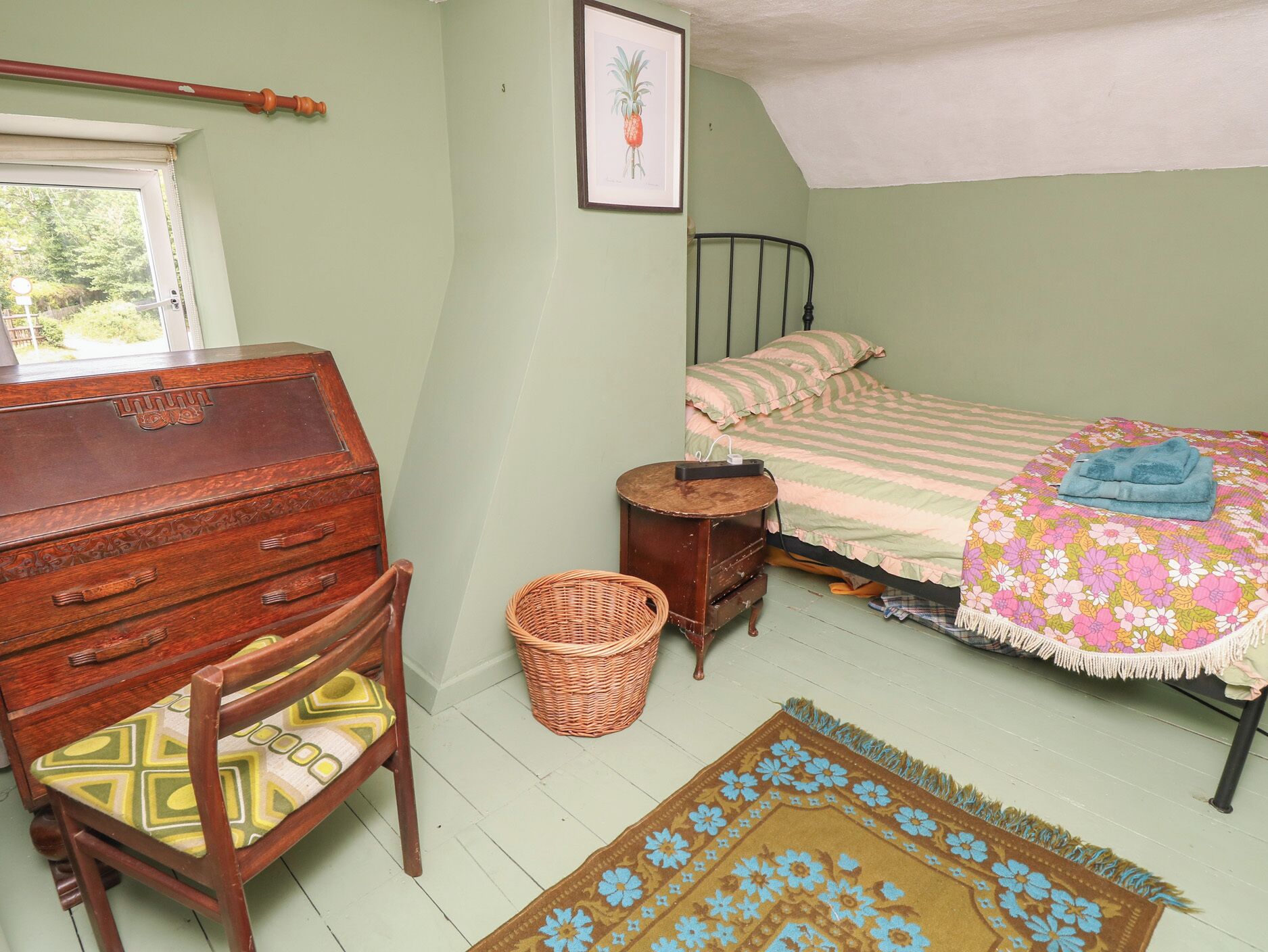 A bedroom with a wooden dresser, a bed with a floral bedspread, a wicker basket, and a small round table.