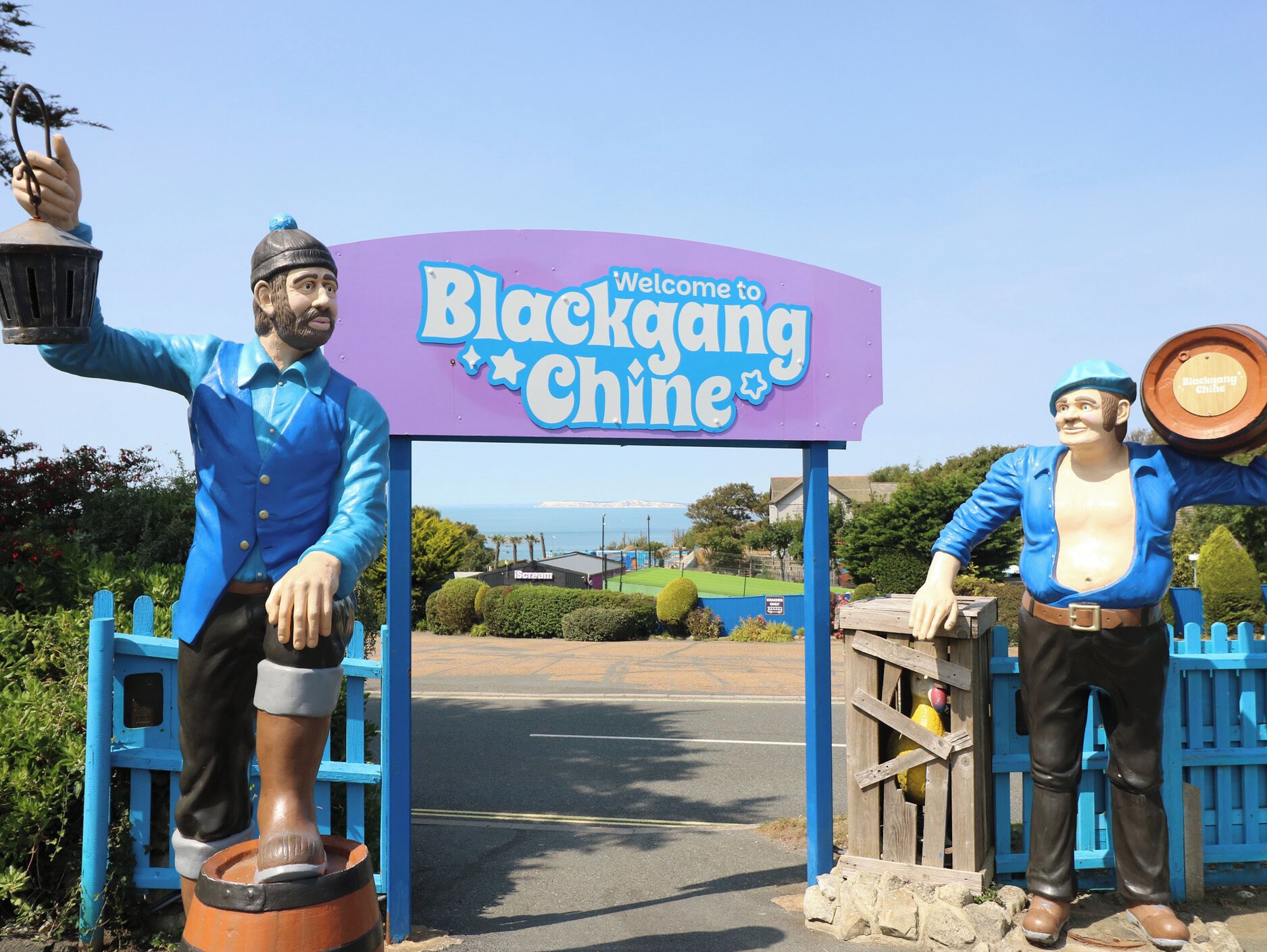 A sign welcoming visitors to Blackgang Chine with two statues of men holding lanterns and a barrel.