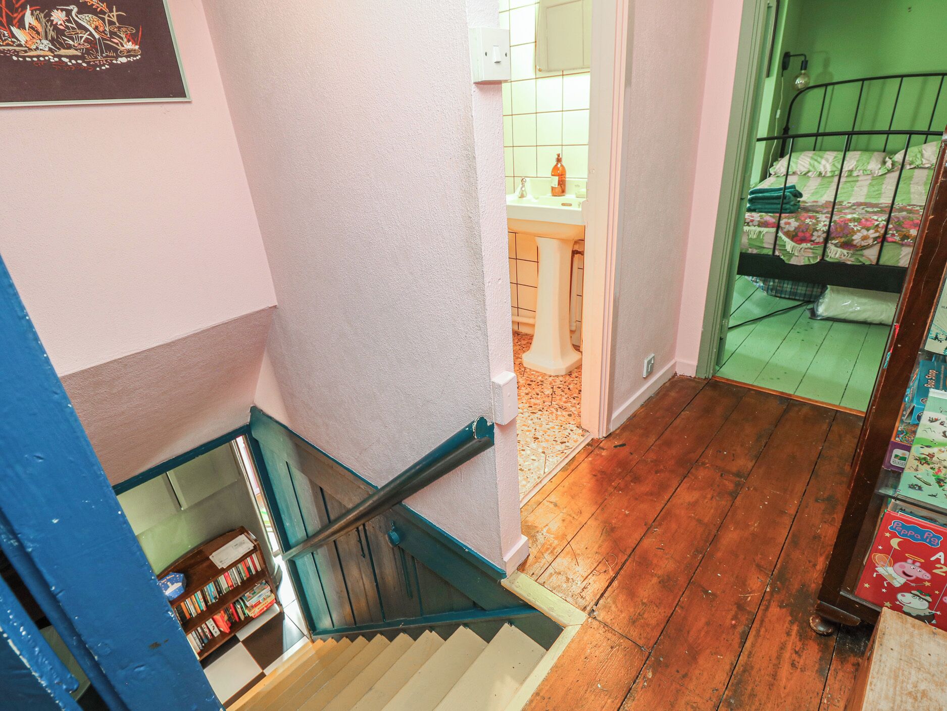 A staircase leading to a room with a green bed, a bathroom with a white pedestal sink, and a bookshelf.