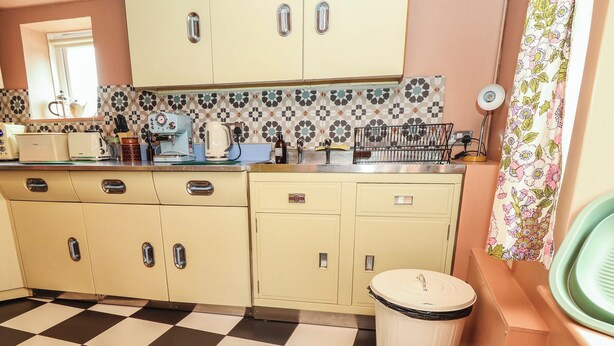 A kitchen with checkered flooring, cream-colored cabinets, a tiled backsplash, and a trash can. - Dairy Cottage (Ventnor)