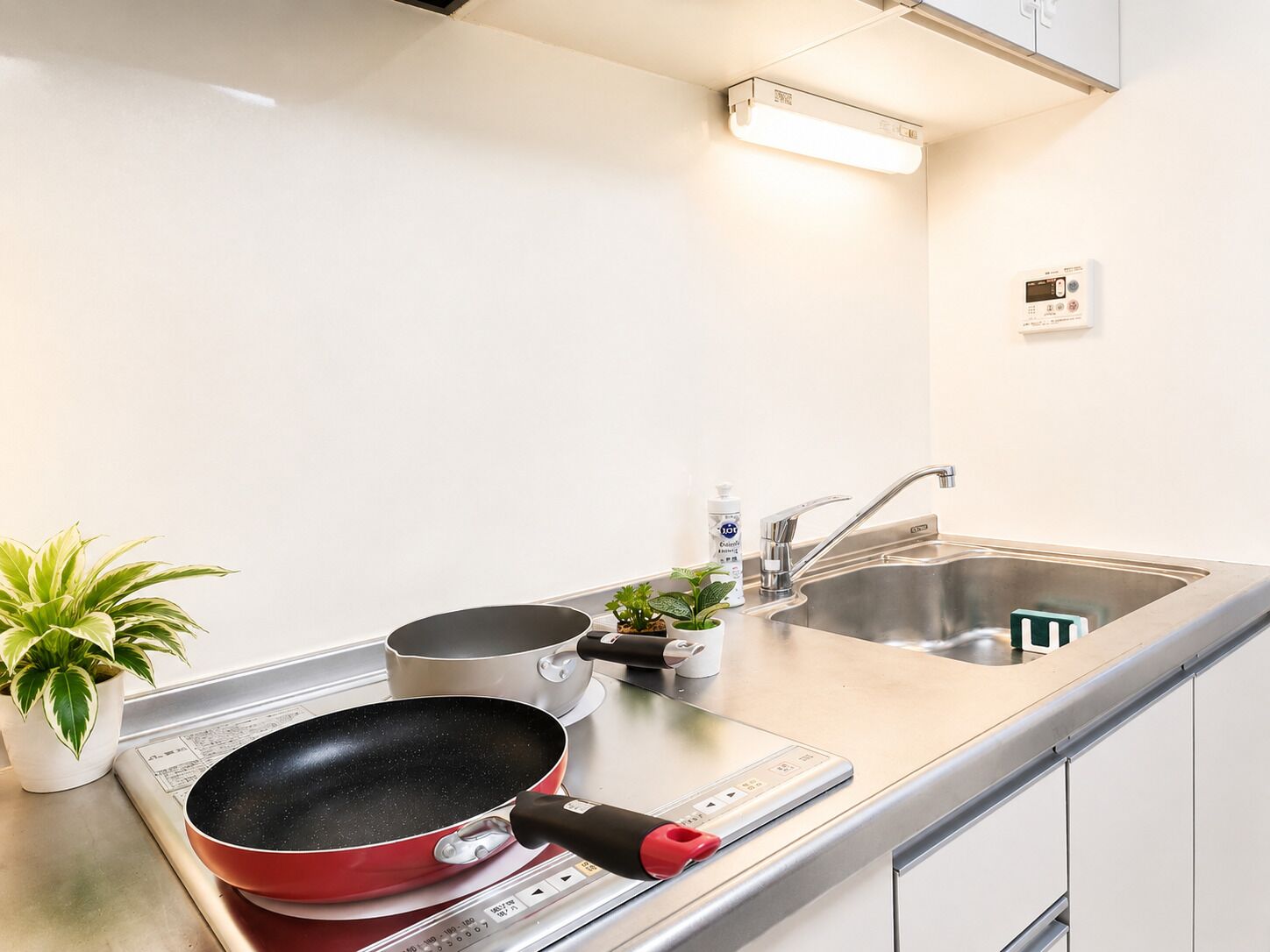 Une cuisine équipée d’une cuisinière, d’un évier, d’une poêle rouge, d’une petite plante en pot et d’un flacon de savon pour les mains.