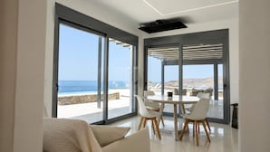 A modern interior with large sliding glass doors, a round dining table, white chairs, and a sofa with a checkered throw. - Kythnos Luxury Villa Aegean View Private Pool Gem (Kithnos)