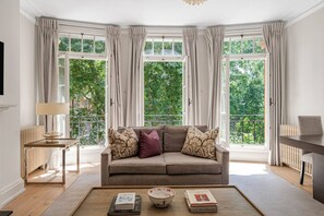 Ein Zimmer mit einer Couch, einem Tisch und Blick auf Grün hinter großen Fenstern.