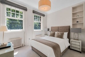Ein Schlafzimmer mit einem großen Bett, zwei Nachttischen, einem Heizkörper und einem Fenster mit Blick auf Bäume.