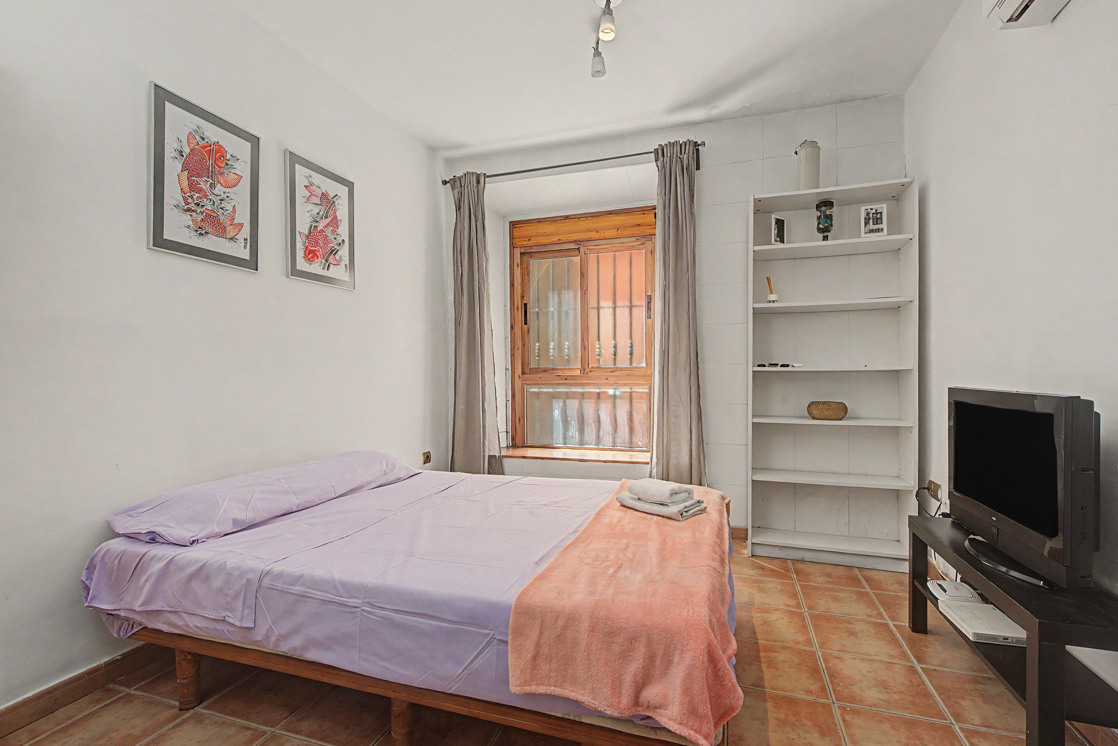 Une chambre à coucher avec un lit, une télévision, une étagère avec des objets et une fenêtre avec des rideaux.