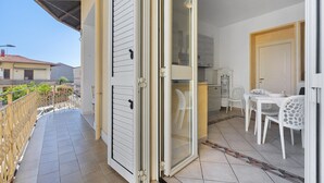 Un balcón con puerta corredera, un comedor con sillas y mesa blancas, y una cocina con armarios y un refrigerador.