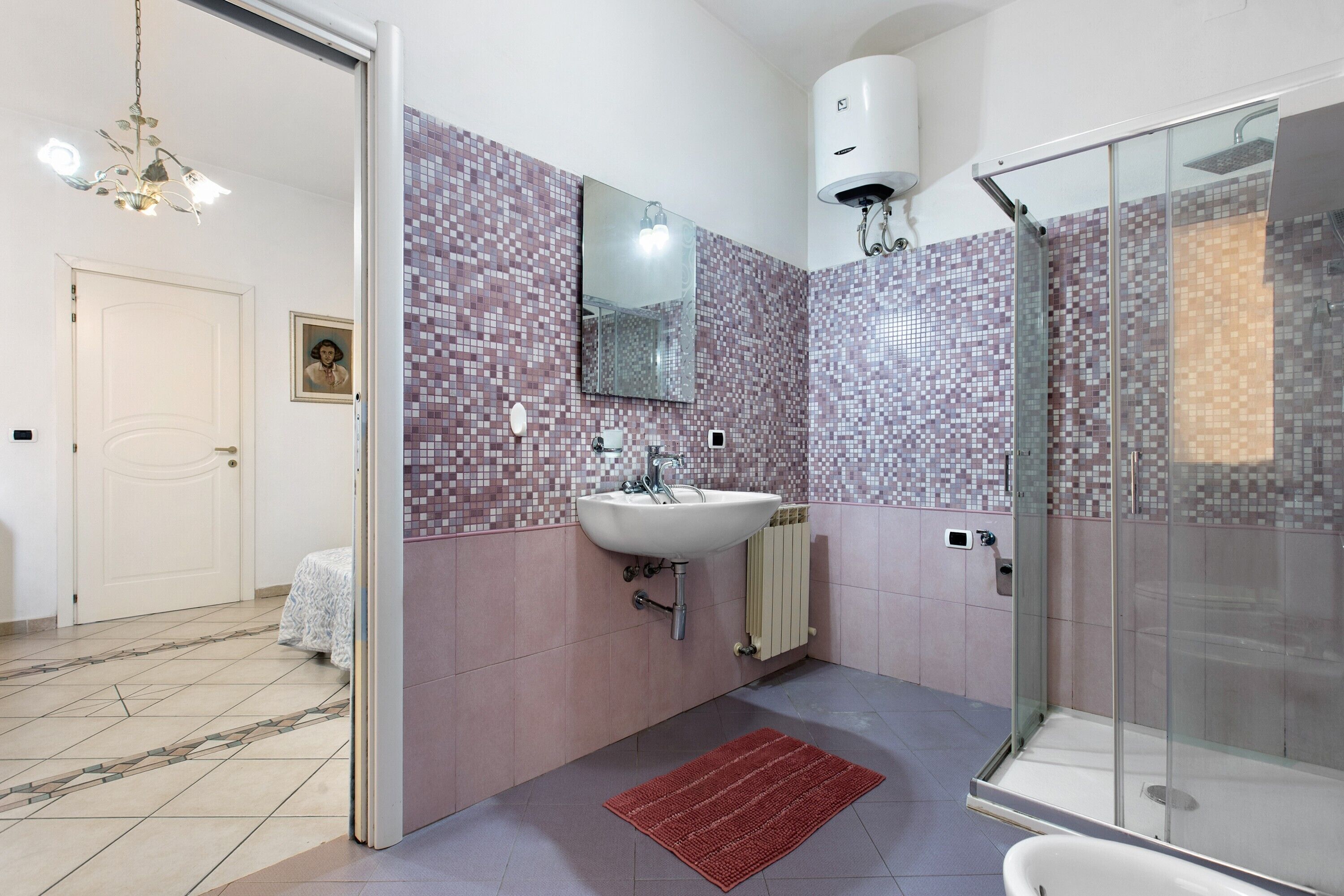 A bathroom with a tiled wall, a sink, a mirror, a shower, and a red rug.