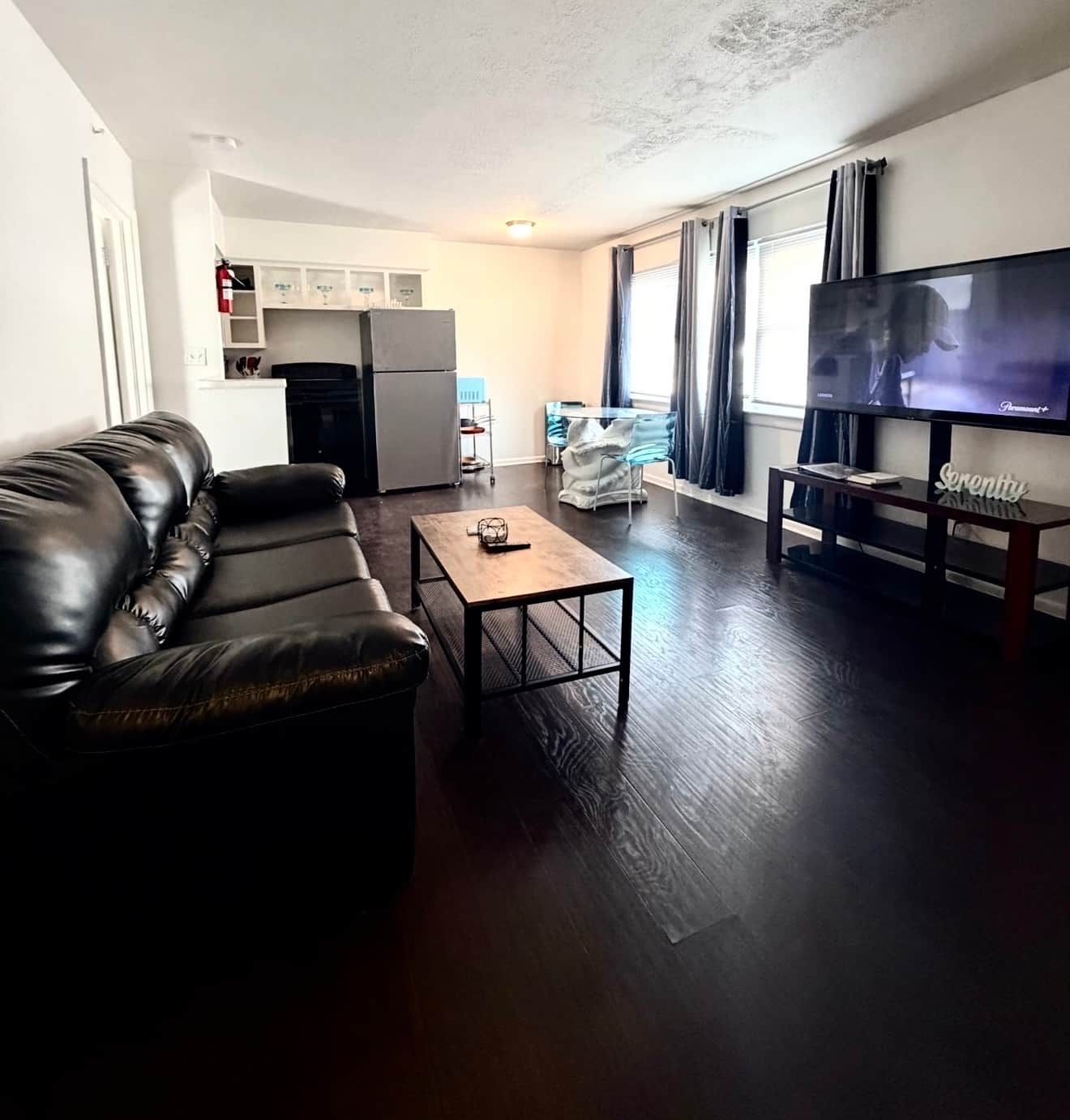 A living room with a leather sofa, a coffee table, a flat-screen TV, and a kitchen area in the background.