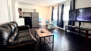 A living room with a leather sofa, a coffee table, a flat-screen TV, and a kitchen area in the background. - The SEA-renity 16 cozy apartment across from the beach (Norfolk)