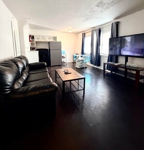 A living room with a leather sofa, a coffee table, a flat-screen TV, and a kitchen area in the background.