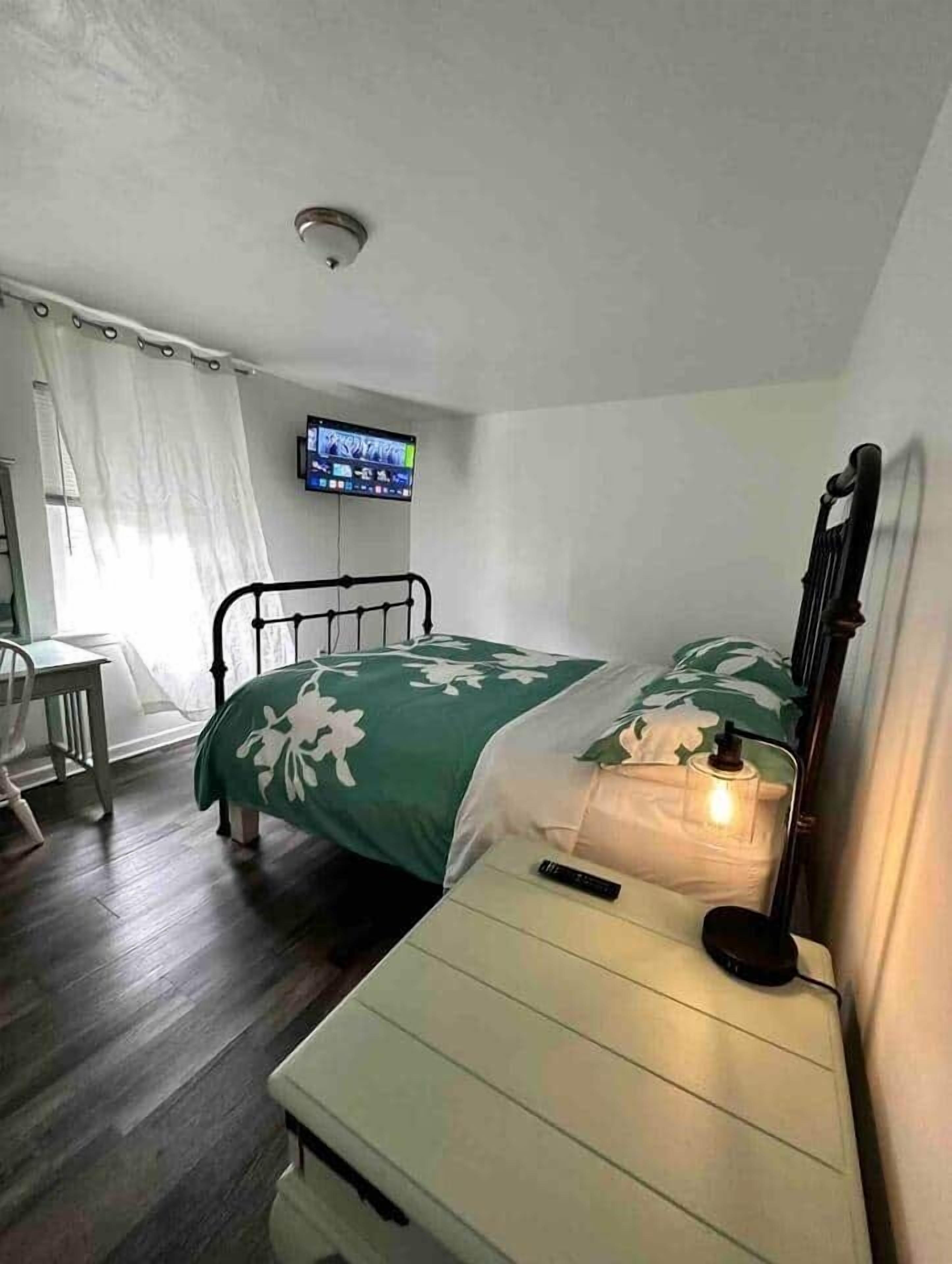 A bedroom with a bed, bedside table, chair, TV, and a window with curtains.