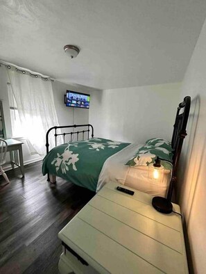 A bedroom with a bed, bedside table, chair, TV, and a window with curtains. - SEA-clusion 7 cozy apartment across from the beach (Norfolk)