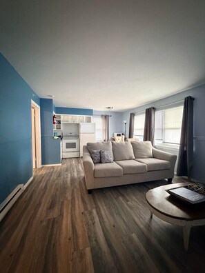 A living room with a beige sofa, a small round table, and a kitchen area with a refrigerator and microwave. - The SEA-renity cozy apartment across from the beach (Norfolk)