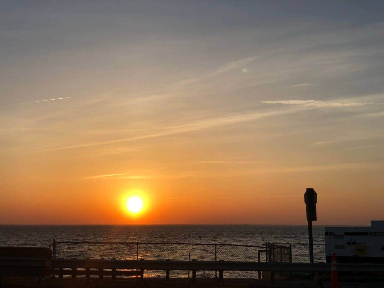 A sunset over the ocean with a fence and a street lamp in the foreground.