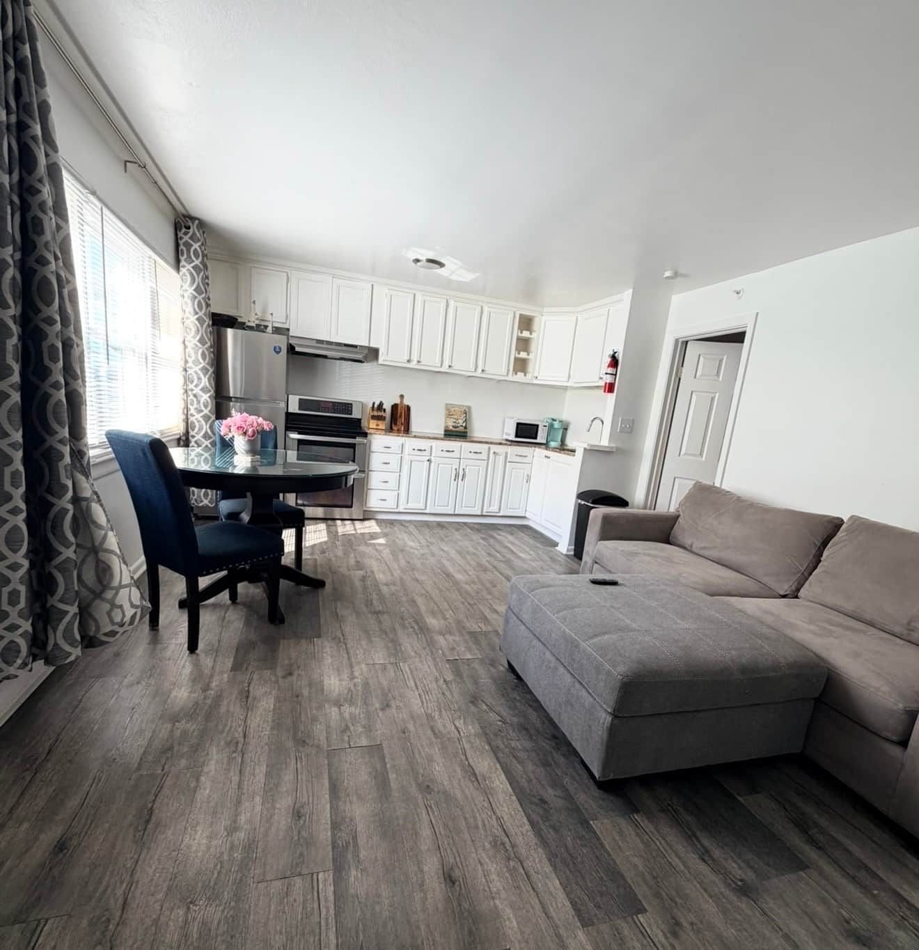 A modern living room with a grey sofa, a dining table with chairs, and a kitchen area with white cabinets and appliances.