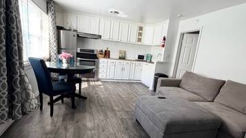 A modern living room with a grey sofa, a dining table with chairs, and a kitchen area with white cabinets and appliances.