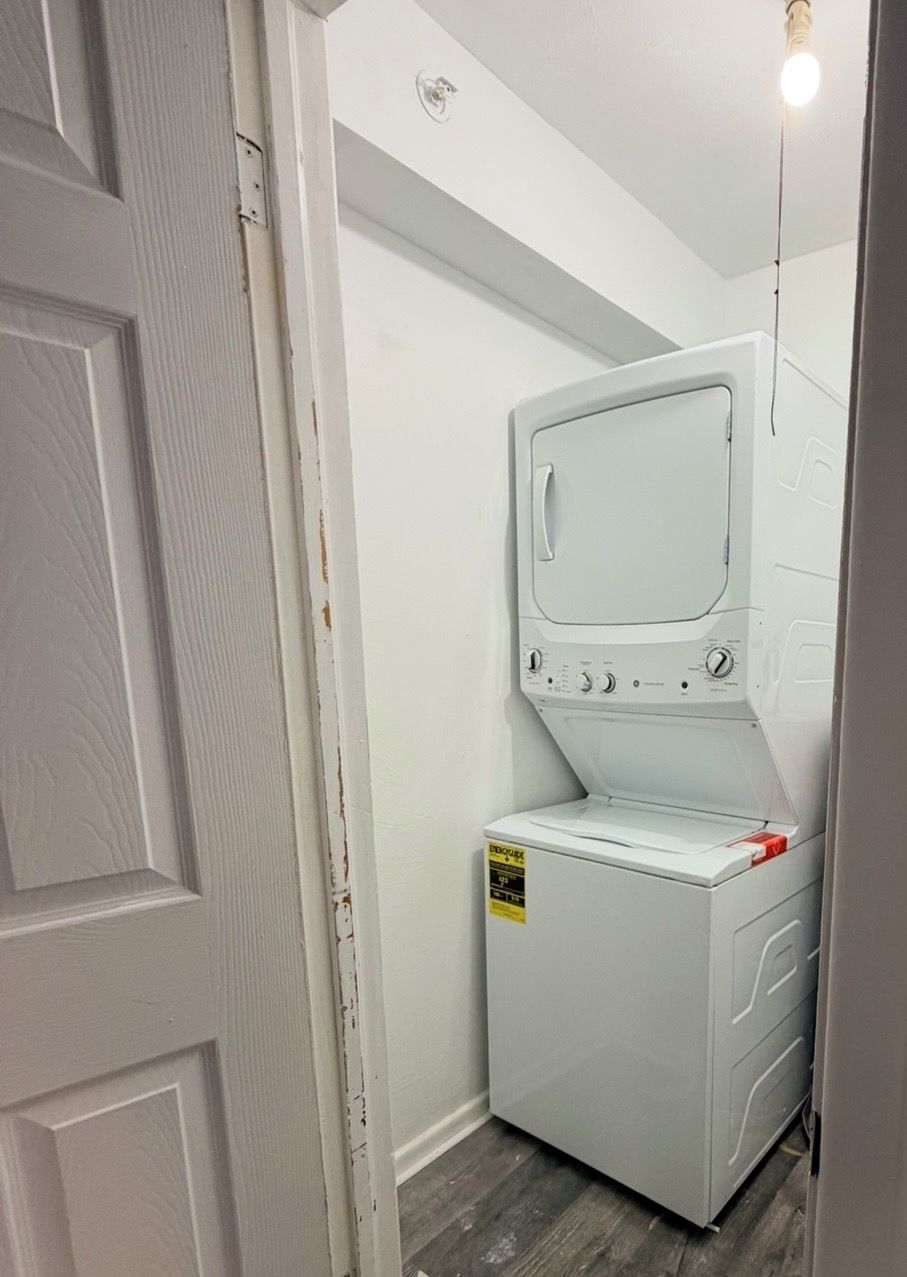 A compact, white laundry room with a built-in washer and dryer, a small shelf, and a light fixture above.
