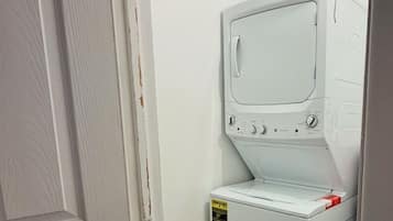 A compact, white laundry room with a built-in washer and dryer, a small shelf, and a light fixture above.