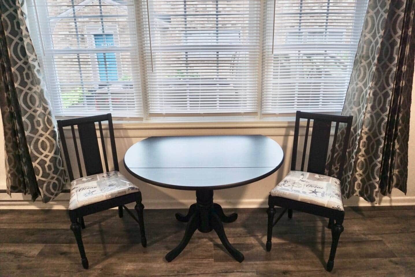 Een ronde tafel met twee stoelen, een raam met zonwering en behang.