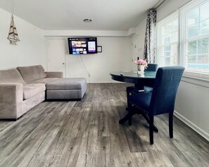 Un salon moderne avec un canapé d’angle gris, une table à manger ronde et une chaise bleue, ainsi qu’un téléviseur à écran plat fixé au mur.