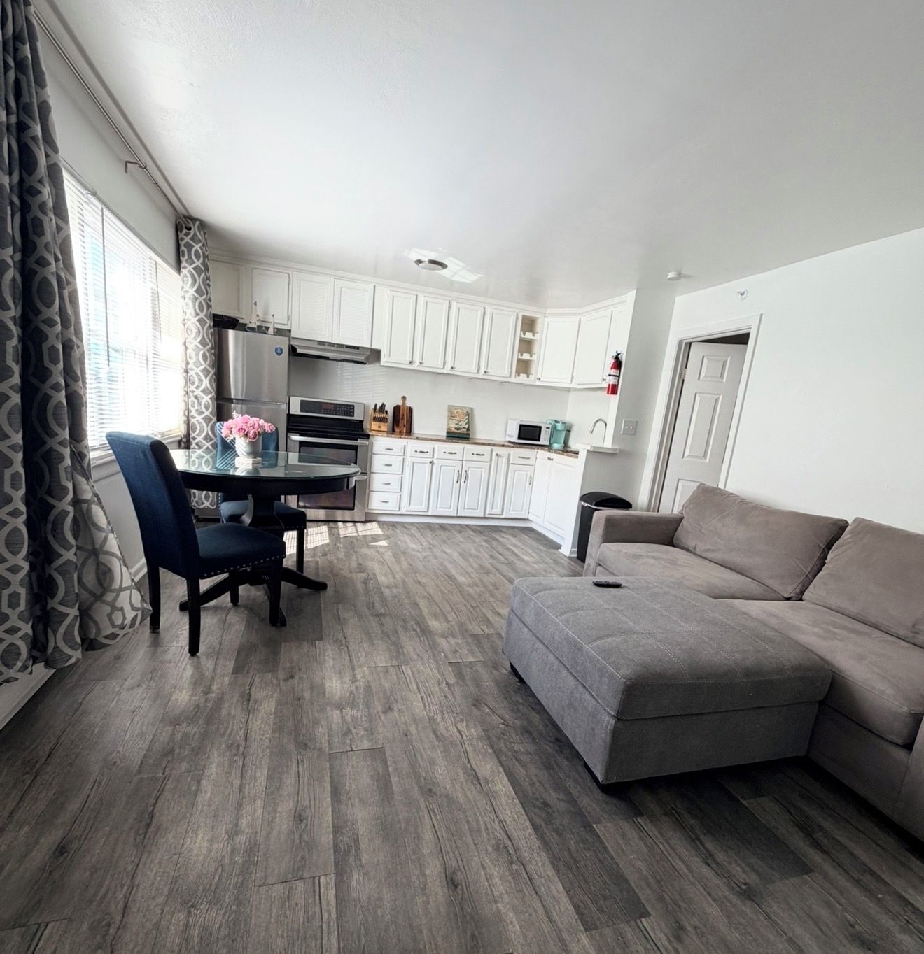 Een moderne woonkamer met een grijze bank, een eettafel met stoelen, en een keuken met witte kasten en een magnetron.