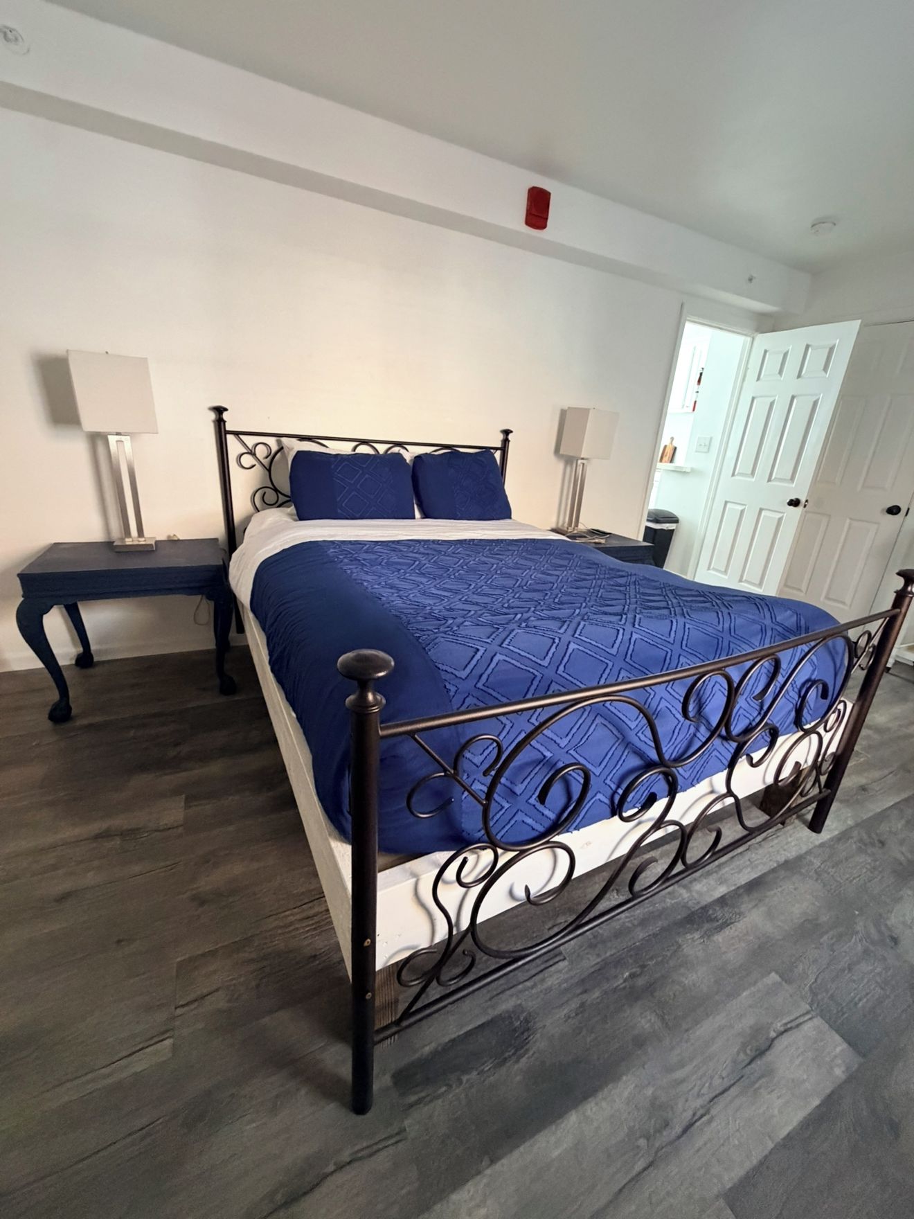 Un dormitorio con estructura de cama metálica, colcha azul, dos mesitas de noche con lámparas, una puerta blanca y suelo de madera.