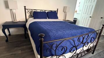 Un dormitorio con estructura de cama metálica, colcha azul, dos mesitas de noche con lámparas, una puerta blanca y suelo de madera.