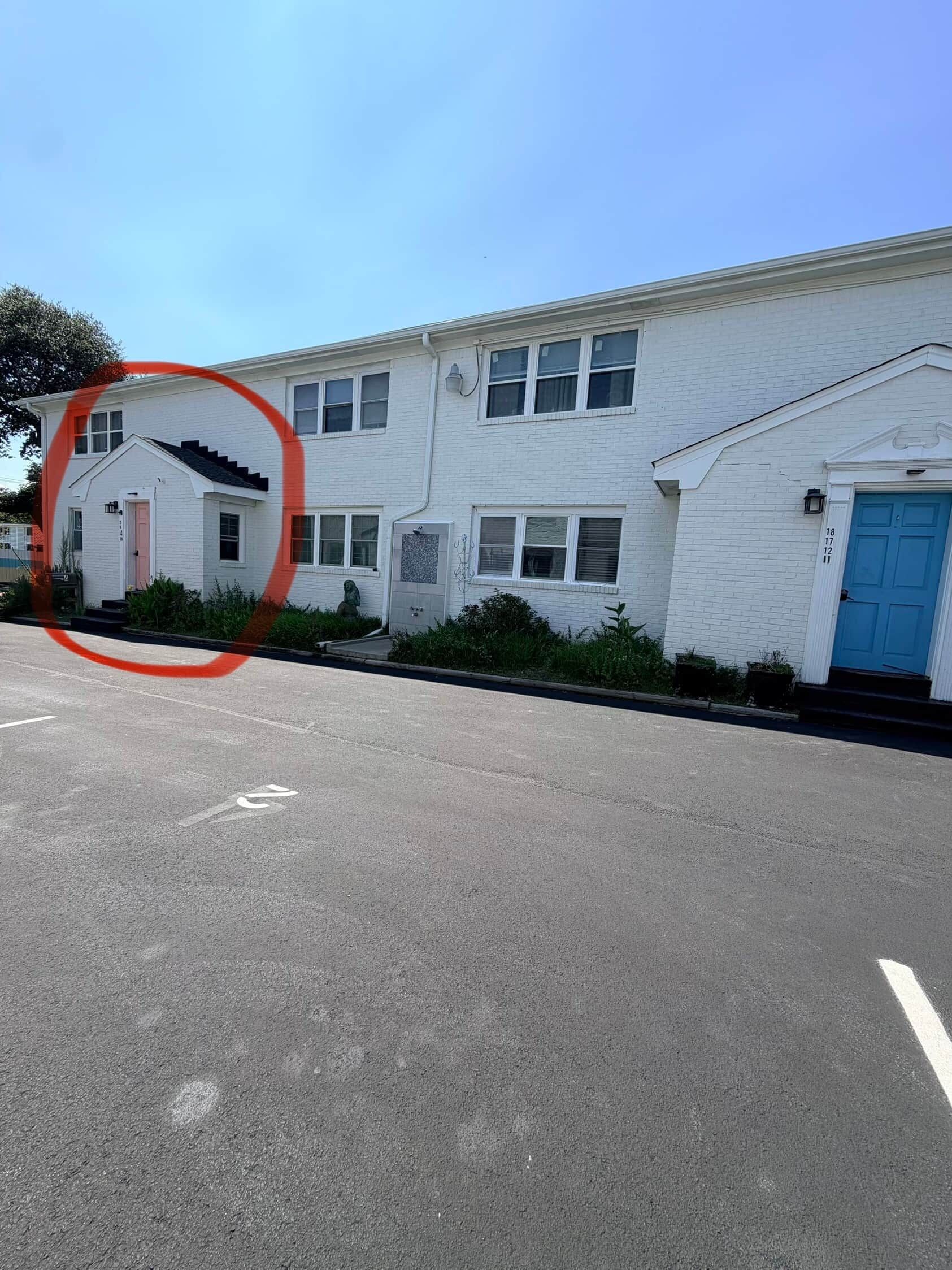 Un bâtiment blanc avec une porte bleue, un petit jardin et un parking.