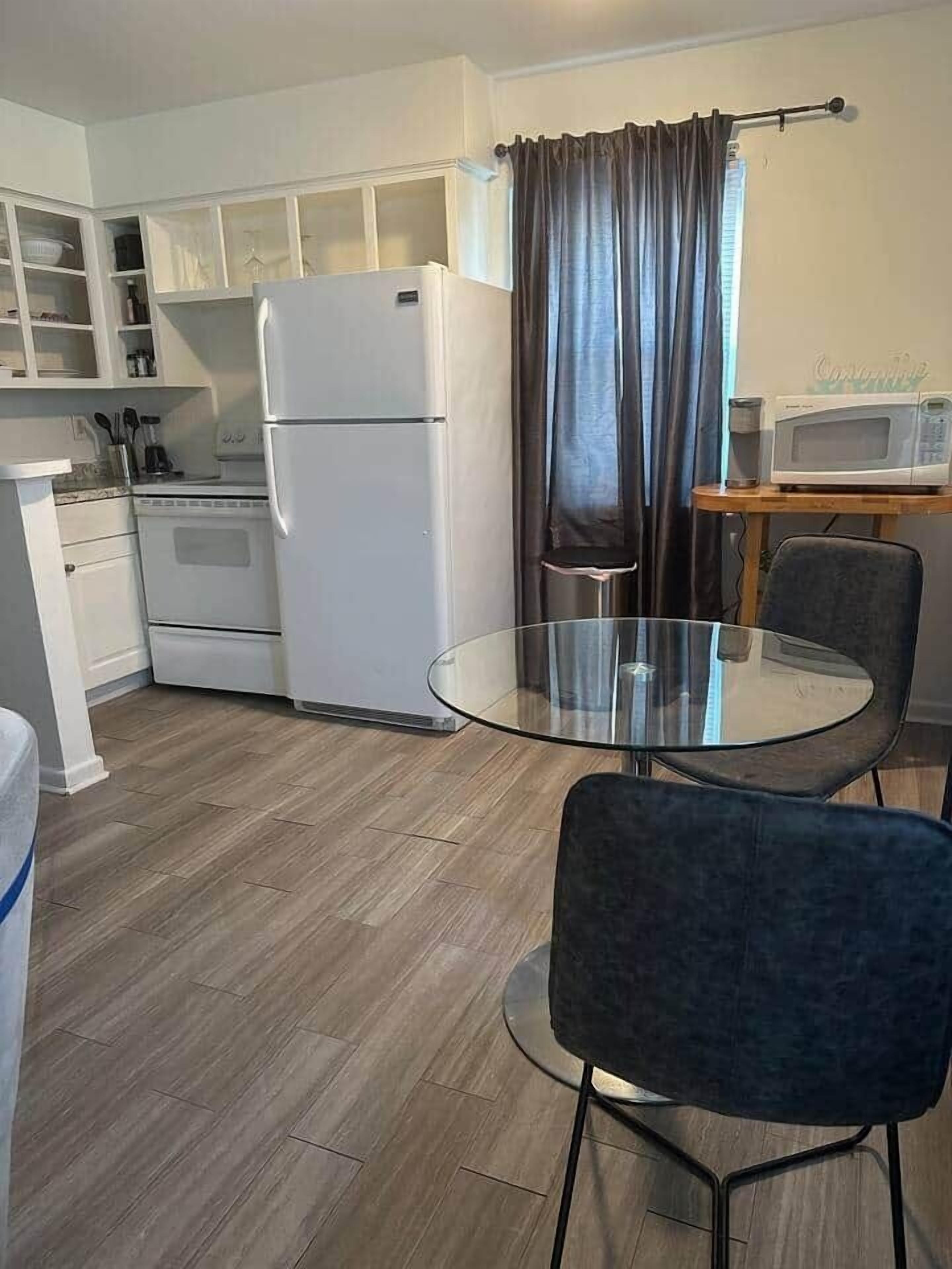 A compact kitchen with white appliances, a glass table, and a dark chair.