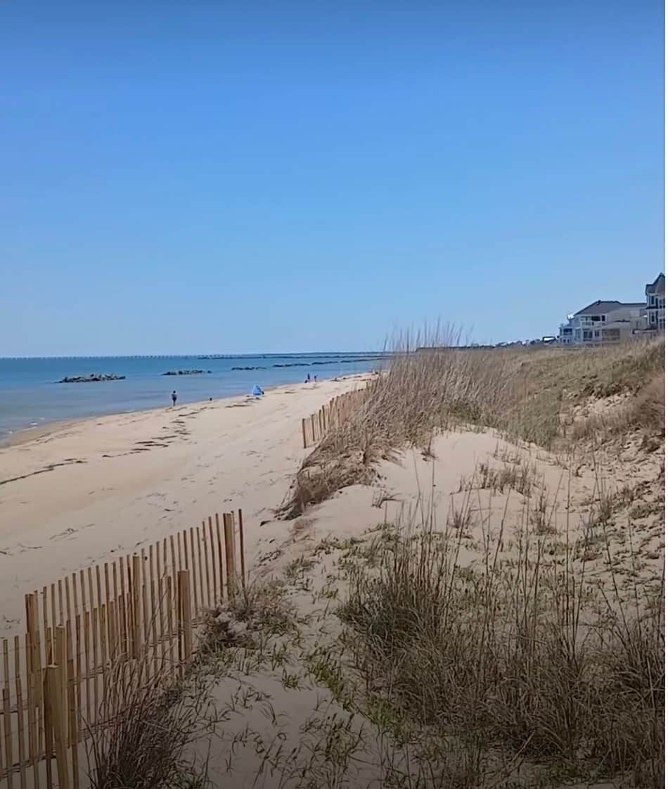 A sandy beach with a wooden fence, grassy dunes, and a clear blue sky.