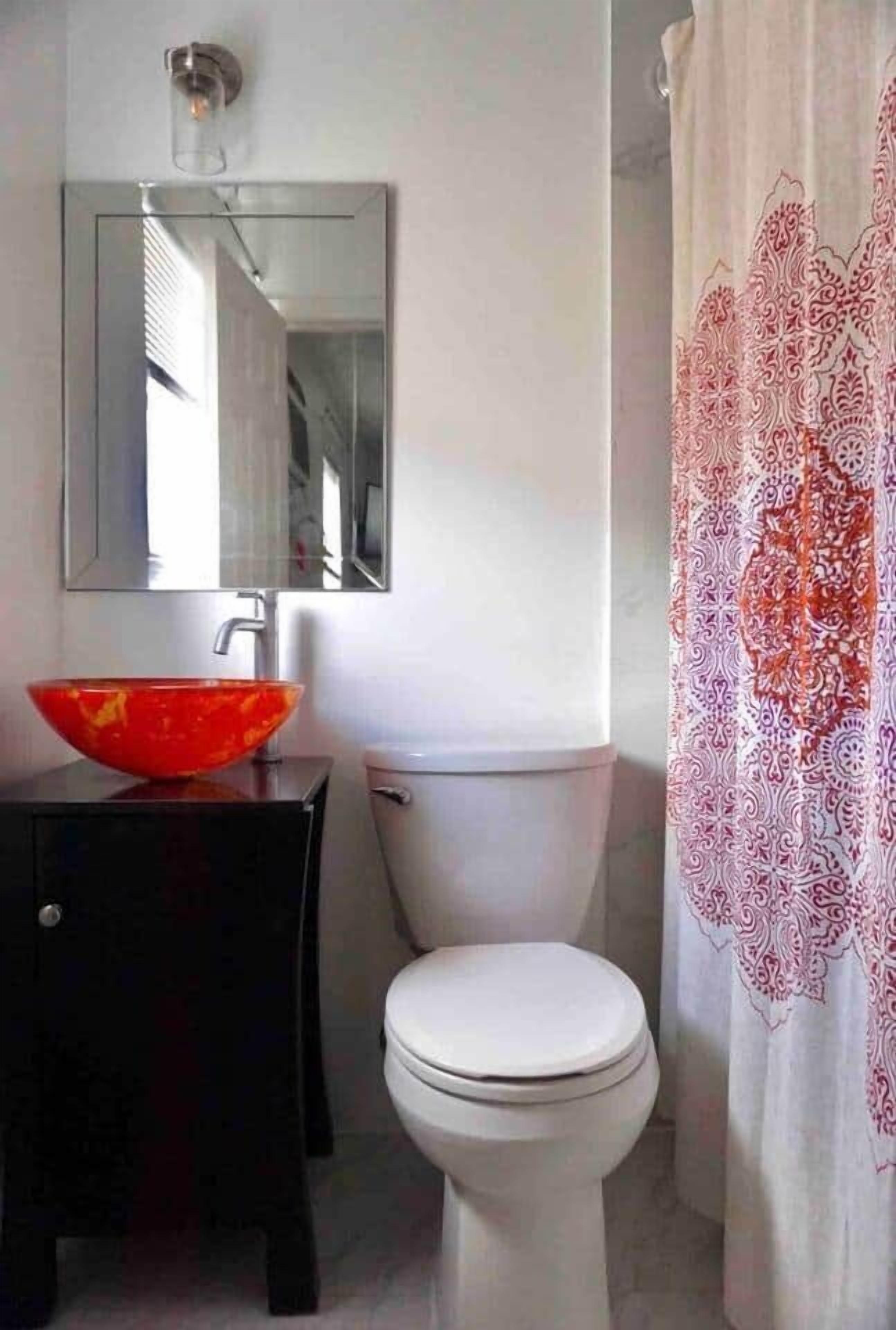A bathroom with a white toilet, a red bowl sink, a black vanity, a mirror, and a patterned shower curtain.
