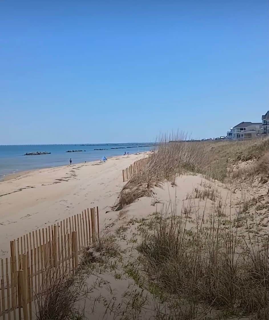 Una playa arenosa con una cerca de madera, dunas cubiertas de hierba y un cielo azul despejado.