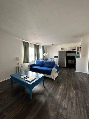 Un espacio habitable compacto con sofá azul, una mesita de centro, una zona de cocina con refrigerador y horno, y una mesa de comedor.