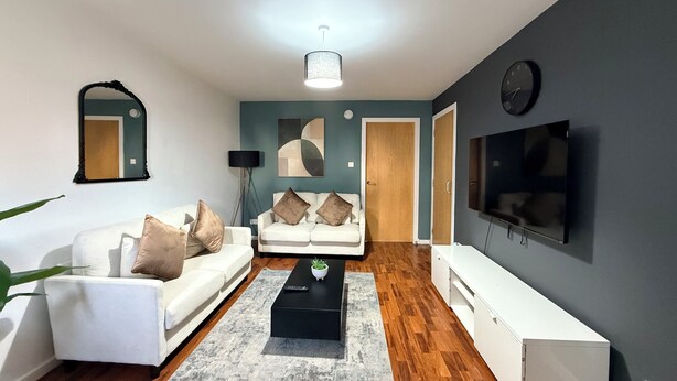 Un salon moderne avec un canapé blanc, une table basse noire, un tapis gris, un téléviseur fixé au mur et un plancher en bois.