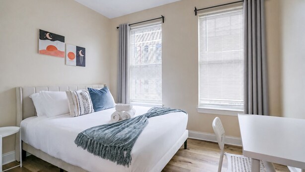 A bedroom with a bed, bedside tables, a desk, and two windows with curtains. - The Pennside 2BR Stay (Philadelphia)
