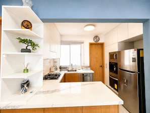 A modern kitchen with white cabinetry, a marble countertop, a built-in oven, and a stainless steel refrigerator.