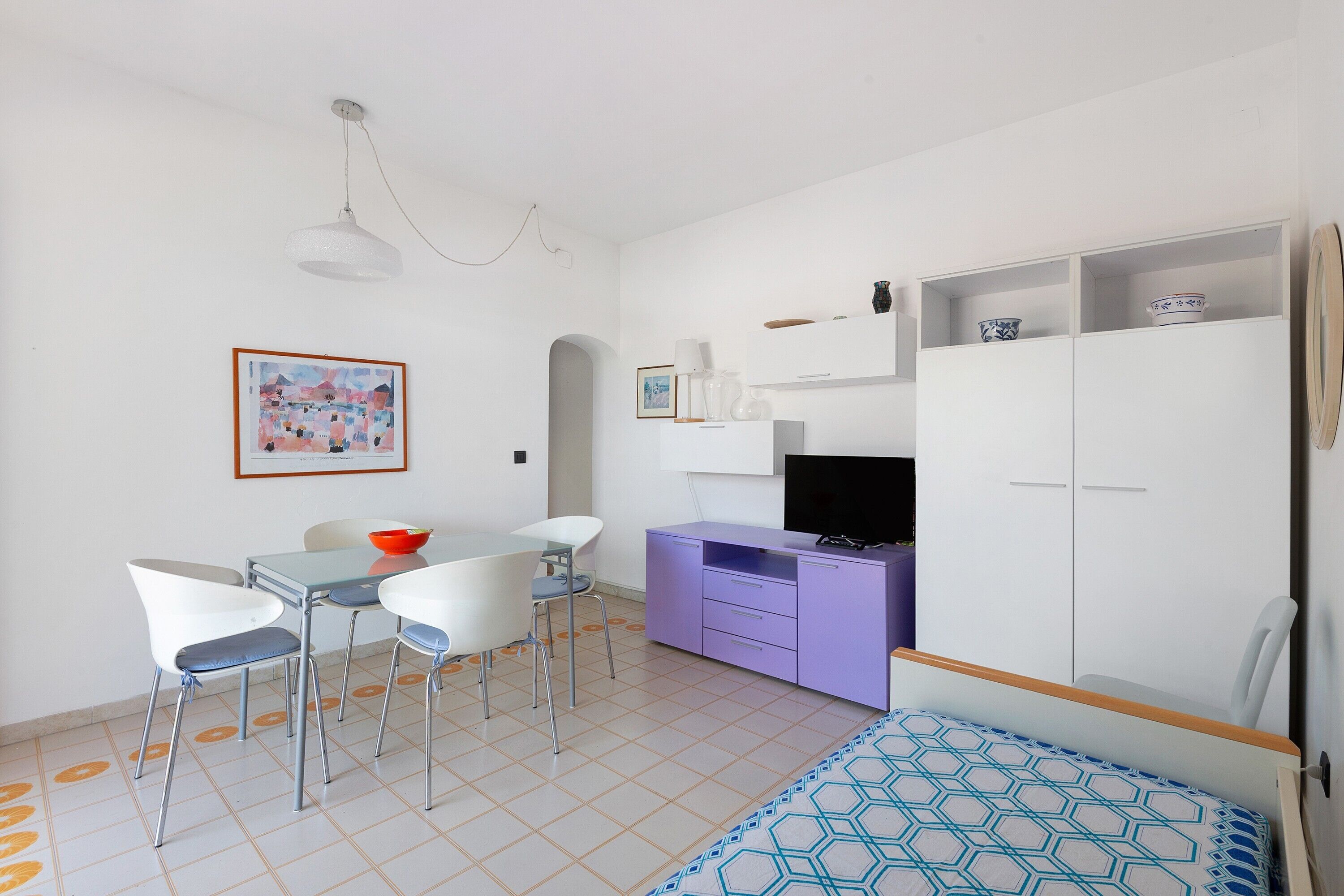 Een compacte kamer met een eettafel, stoelen, een paars kastje, een televisie en een bed met een decoratieve dekbedovertrek.