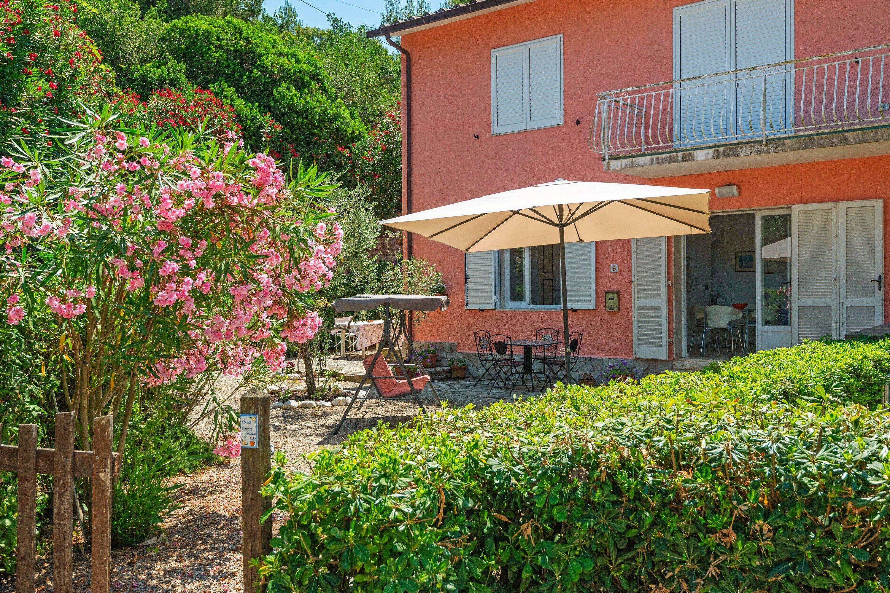 Una casa de dos pisos con un balcón, un patio con mesa y sillas, y un jardín lleno de flores rosadas.