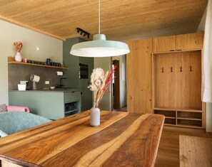 A wooden dining table with a vase of dried flowers, a hanging light fixture, and a kitchen area with a green countertop and wooden cabinets.