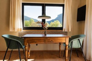 A dining area with a wooden table, two chairs, a vase with dried flowers, and a view of mountains through a large window.